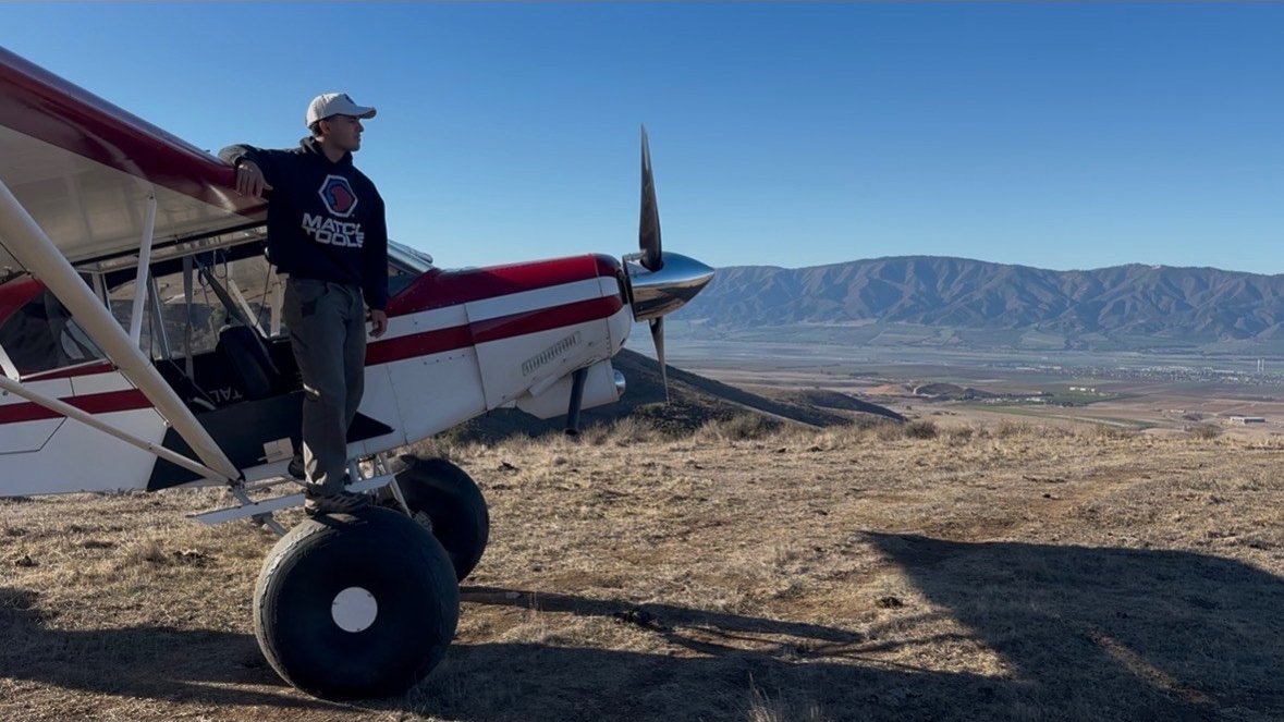 Gabriel standing on the tire of a backcountry taildragger on a mountaintop above the valley behind him.