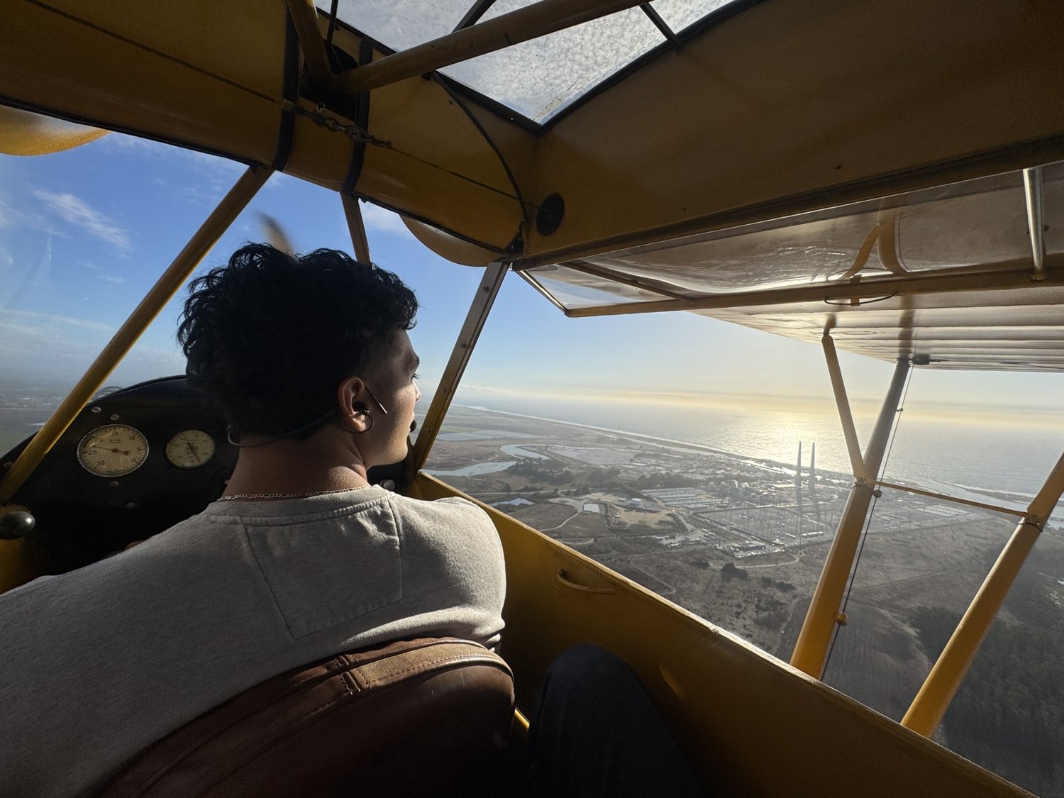 Gabriel flying an open-cockpit biplane over the Monterey Bay coastline at sunset.
