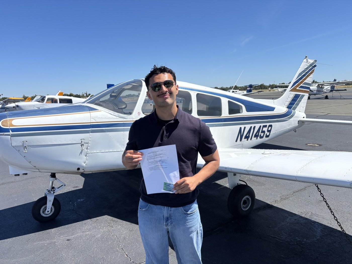 Gabriel in sunglasses holding his certificate in front of the Piper Warrior N41459 on a sunny ramp.