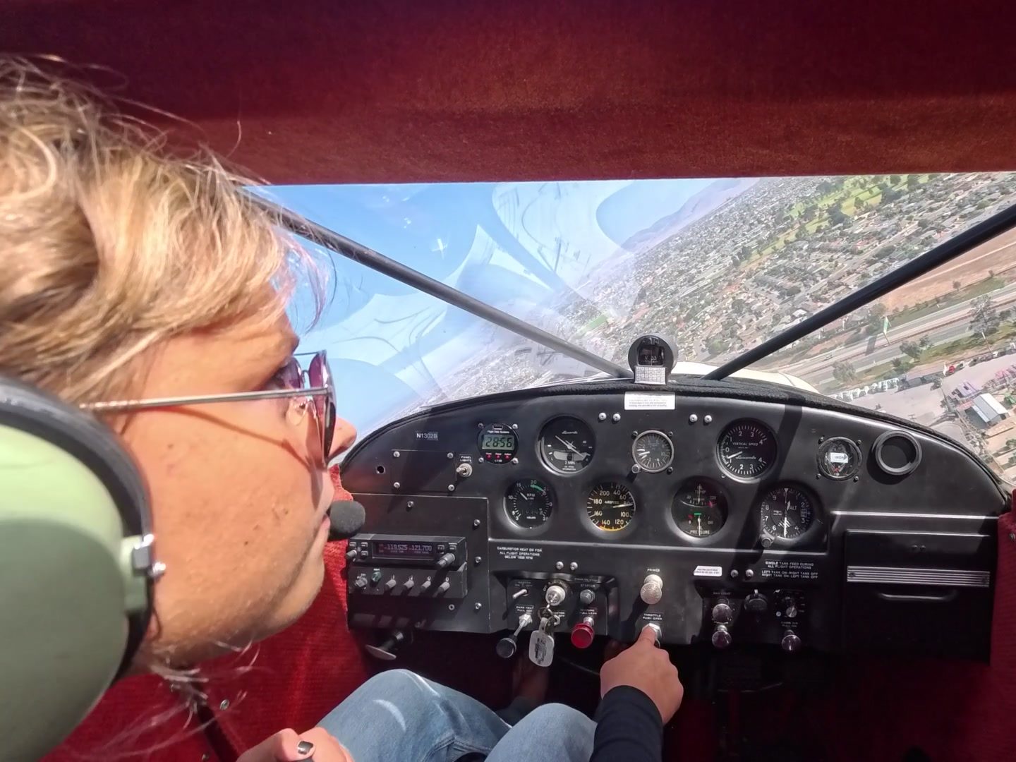 Hadley at the controls of the Luscombe in the traffic pattern, wearing a headset and sunglasses, with Salinas below.