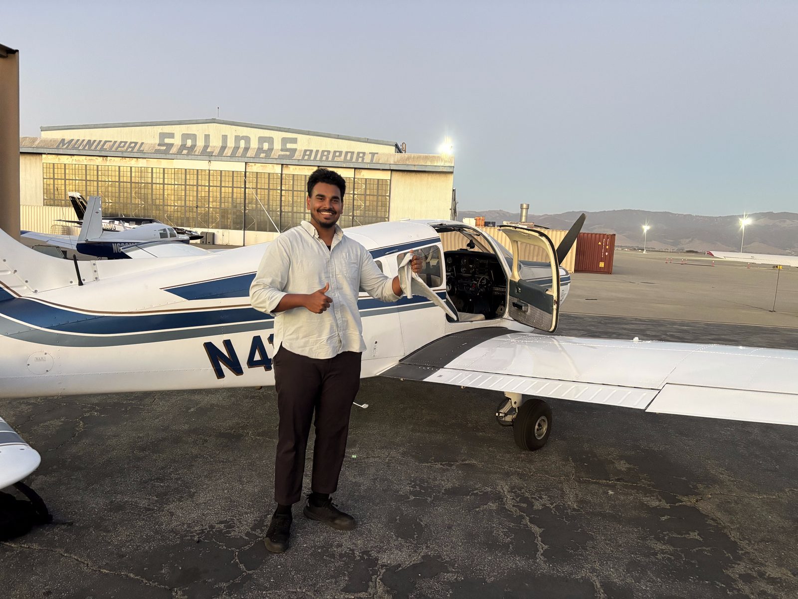 Nazeem holding his cut shirt tail and giving a thumbs up in front of the Warrior, with the Salinas Municipal Airport hangar behind him.